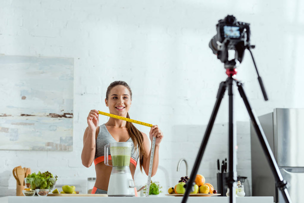 selective focus of cheerful sportswoman holding measuring tape near fruits and digital camera  - Photo, Image