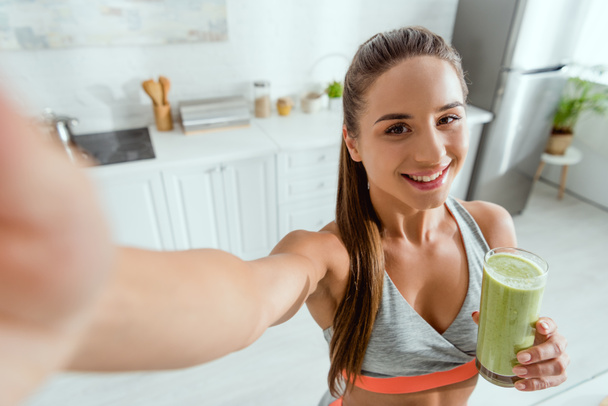 selective focus of happy girl holding green smoothie and looking at camera  - Fotografie, Obrázek