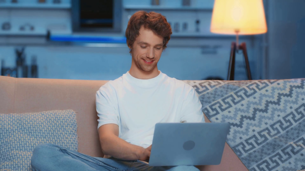 uśmiechnięty człowiek wpisując na laptopie, siedząc na kanapie - Materiał filmowy, wideo
