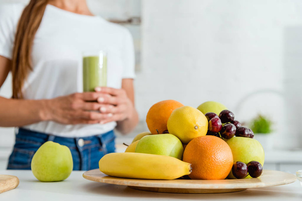 enfoque selectivo de frutas deliciosas maduras cerca de niña sosteniendo vidrio con batido verde
  - Foto, Imagen