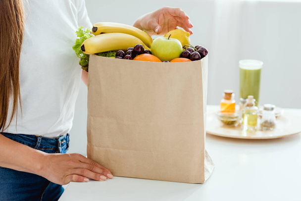 cropped view of young woman standing near paper bag with fruits  - 写真・画像