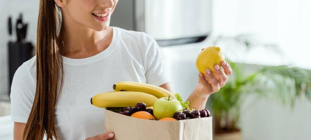 panoramic shot of positive  woman looking at lemon near paper bag with fruits  - Photo, Image