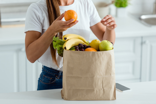 cropped view of woman with orange near paper bag and smartphone with blank screen - Foto, Bild