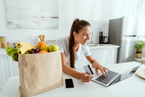 szczęśliwy dziewczyna przy pomocy laptop chwila online zakupy w pobliżu papier worek rezygnować owoce   - Zdjęcie, obraz