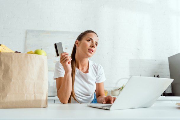 zamyślony dziewczyna trzyma karty kredytowej podczas korzystania z laptopa w pobliżu torby papierowej  - Zdjęcie, obraz