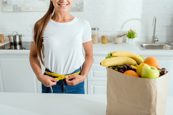 обрезанный вид веселой молодой женщины, измеряющей талию возле бумажного пакета с продуктами
  - Фото, изображение