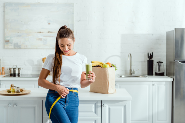 happy young woman measuring waist and holding glass with smoothie near groceries  - Foto, afbeelding