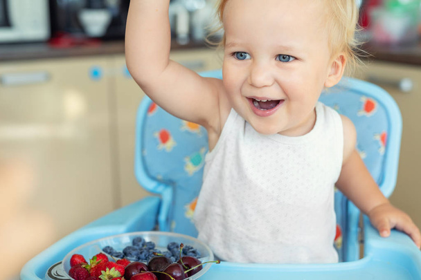 Очаровательный милый белый блондин-мальчик наслаждается вкусом различных сезонных свежих зрелых органических ягод, сидя в высоком кресле на домашней кухне. Счастливый малыш делится натуральной сладкой здоровой пищей с другом
 - Фото, изображение