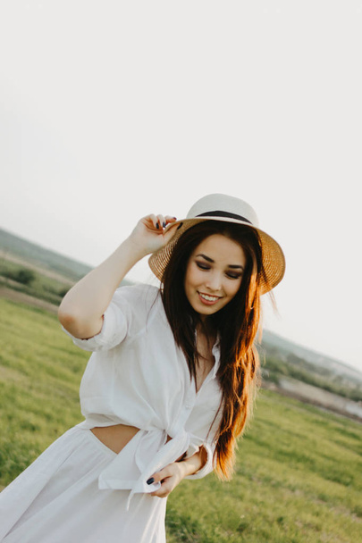Portrait of charming carefree long hair asian girl in white clothes and straw hat enjoys life in field at sunset. Sensitivity to nature concept - Photo, Image