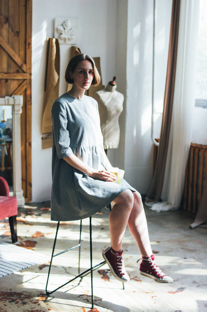 Young woman in simple grey dress with yellow cup sitting on chair in art work studio, full length portrait - Foto, imagen