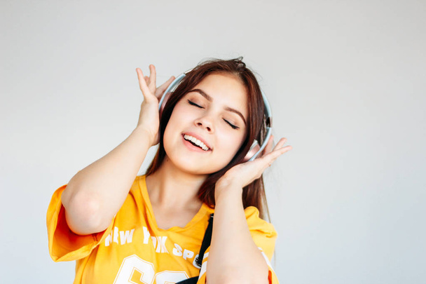 Счастливая беззаботная улыбающаяся девочка-подросток с темными длинными волосами в желтой футболке слушающая музыку в наушниках, изолированных на сером фоне
 - Фото, изображение