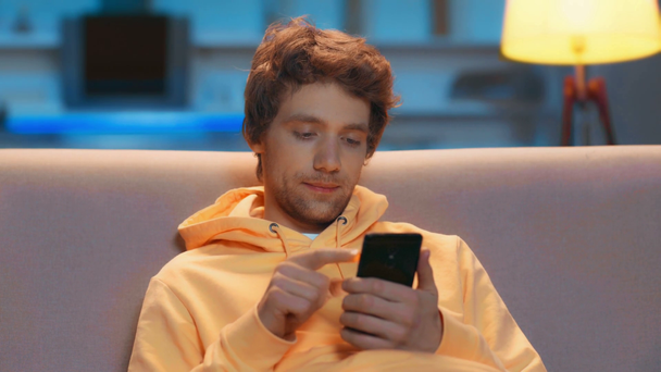 srác ül a kanapén, és okostelefont használ a nappaliban - Felvétel, videó