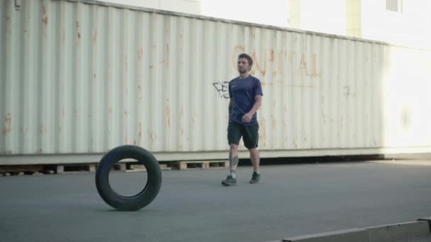 Young bearded male do exercises with car tire outdoors, industrial or urban background - 映像、動画