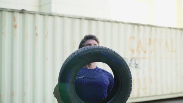 Young bearded male do exercises with car tire outdoors, industrial or urban background - Кадры, видео