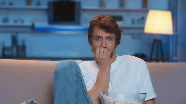scared man eating popcorn while watching horror film on sofa at night - Filmagem, Vídeo