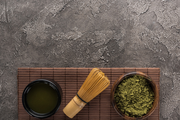 Ansicht von grünem Matcha-Tee mit Schneebesen auf Bambusmatte auf dunklem Steintisch - Foto, Bild