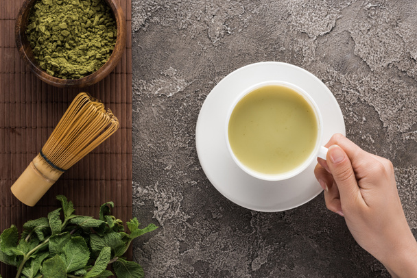 cropped view of woman holding matcha matcha tea on table with whisk, powder and mint  - Valokuva, kuva