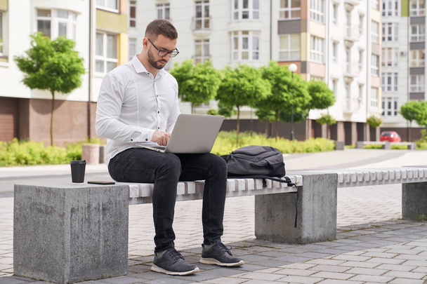 Handsome man sitting on bench, working on laptop outdoors. - Valokuva, kuva