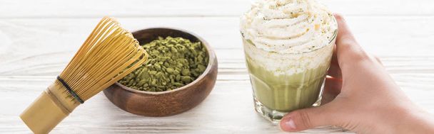 Cropped view of woman holding matcha matcha tea with whipped cream on table with whisk and powder  - Photo, Image