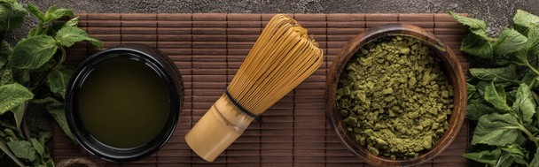 Ansicht von grünem Matcha-Teepulver mit Minze und Schneebesen auf Bambusmatte auf dunklem Steintisch - Foto, Bild