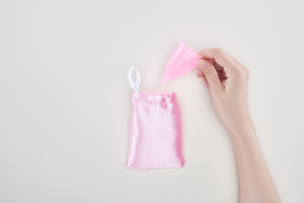 cropped view of woman holding pink plastic menstrual cup near bag isolated on grey - Foto, imagen