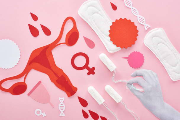 vista recortada de mano blanca sosteniendo tampón sobre fondo rosa con toallas sanitarias, papel cortado órganos internos reproductores femeninos y gotas de sangre
 - Foto, Imagen
