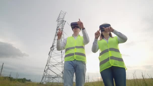 Um homem e uma mulher engenheira em óculos VR controlam a distribuição de energia das redes elétricas e a entrega de eletricidade no fundo de torres elétricas com cabos de alta tensão
. - Filmagem, Vídeo