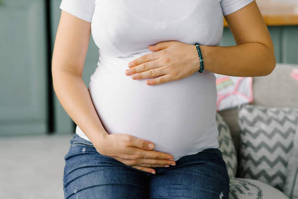 ジーンズと白いTシャツを着た妊娠中の女性が、大きなお腹に手を当てている。赤ちゃんを待っている - 写真・画像