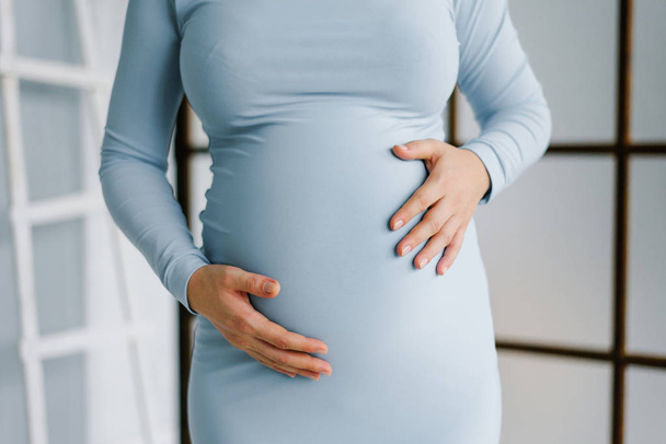 Těhotná žena v pevně modrých šatech jí drží v žaludku její ruce v očekávání narození dítěte a porodu - Fotografie, Obrázek