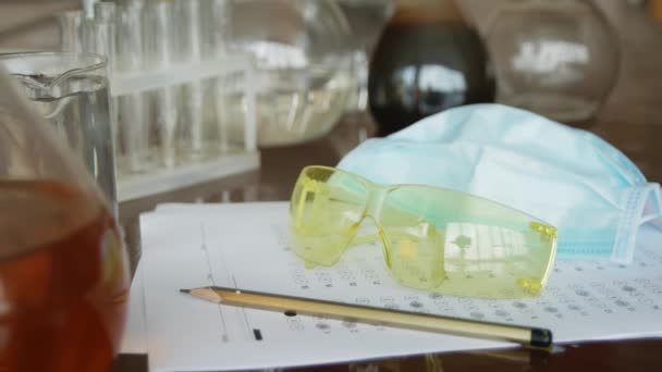 Στην τάξη της χημείας στο τραπέζι είναι τεστ εξετάσεων, μολύβι, γυαλιά και μάσκα για τους μαθητές. Κοντινό, χημικός εξοπλισμός, εκπαιδευτική ιδέα. - Πλάνα, βίντεο
