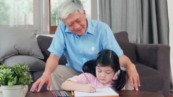 Der asiatische Großvater unterrichtet seine Enkelin beim Zeichnen und Hausaufgaben machen. Senior Chinese, Opa glücklich entspannen mit jungen Mädchen auf dem Sofa im Wohnzimmer zu Hause Konzept liegen. Zeitlupe. - Filmmaterial, Video
