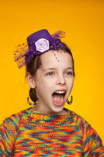 Portrait of a cheerful 8-year-old girl in a miniature hat and in a multi-colored sweater. Yellow background - Fotoğraf, Görsel