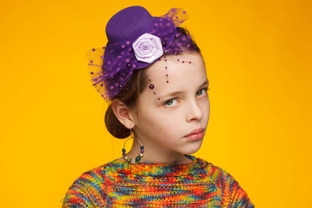 Portrait of a cheerful 8-year-old girl in a miniature hat and in a multi-colored sweater. Yellow background - Photo, image