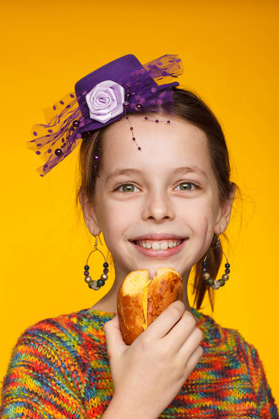 Portrait of a cheerful 8-year-old girl in a miniature hat, a multi-colored sweater and with a sandwich in hand - Photo, image