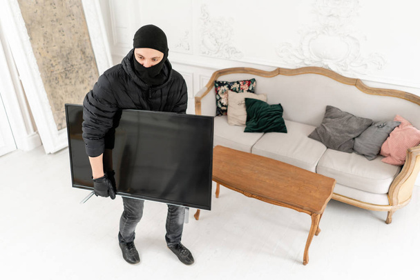 Thief with black balaclava stealing modern expensive television. Luxury apartment with stucco. Man burglar stealing tv set from house - Photo, image