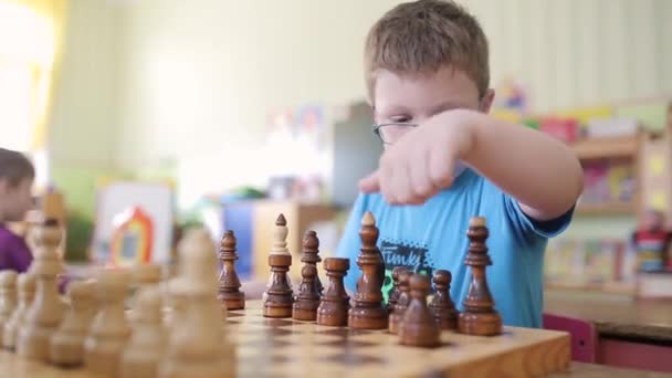 A boy with glasses plays chess. He is sitting at the table playing and smiling emotionally. He wants to checkmate and win. - Imágenes, Vídeo
