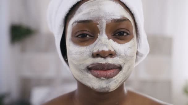 Attractive afro-american girl with white towel on head having a cleansing mask on face looking at camera - Imágenes, Vídeo
