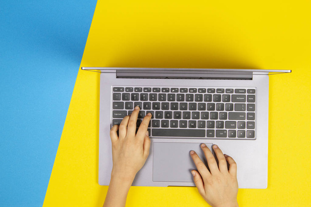 Ręce dzieciak pisania na klawiaturze komputera laptop, widok z góry, żółte i niebieskie tło - Zdjęcie, obraz
