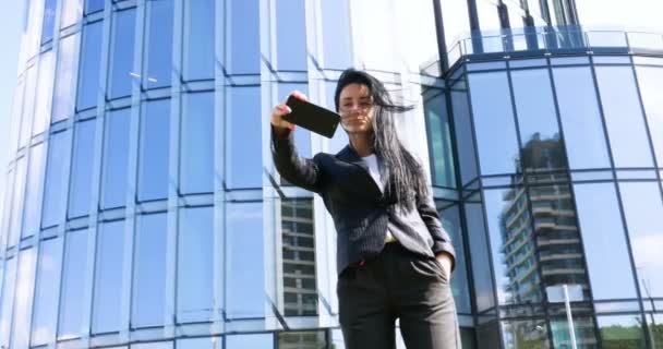Businesswoman making selfies in front of office building. Smiling lady taking photos for her business partners abroad. Concept of: Lifestyle, Mobile, Selfie, Alone, Summer. - Filmagem, Vídeo
