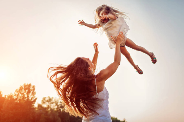 Γυναίκα που παίζει και διασκεδάζει με την κόρη του στο καλοκαιρινό πάρκο το ηλιοβασίλεμα. Μητέρα πετώντας κορίτσι σε εξωτερικούς χώρους. - Φωτογραφία, εικόνα