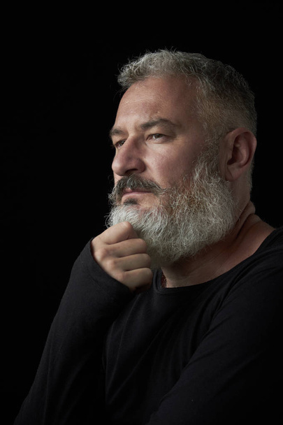 Retrato de un hombre de pelo gris brutal con una barba exuberante gris y una cara estricta sobre un fondo negro, enfoque selectivo
 - Foto, Imagen