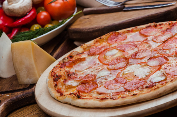 salami pizza on a wooden board, wooden background decorated with vegetables and cutlery - Foto, Imagen