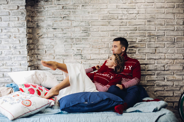 Couple in red Christmas jumpers kissing surrounded by pillows. - Photo, Image
