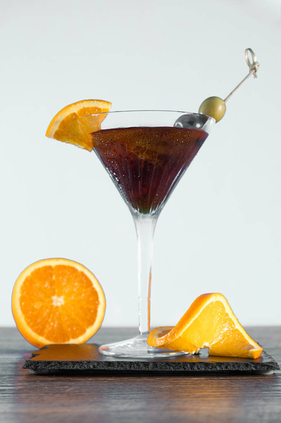 グラスに濃いオレンジの飲み物。アイスキューブとレモンスライス。緑と黒のオリーブヴェルマスとジン. - 写真・画像