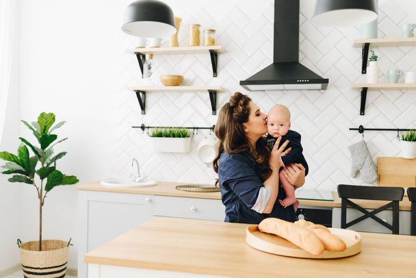 Uma bela jovem mãe sentada na mesa da cozinha de madeira e beijando seu bebê
  - Foto, Imagem