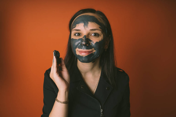 A young woman inflicts a black mask on the face on an orange background. The concept of healthy lifestyle, beauty, body care. - Фото, зображення