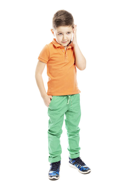 A cute school-age boy in a bright orange polo t-shirt is talking on a smartphone. Full height. Isolated on a white background. - Fotografie, Obrázek