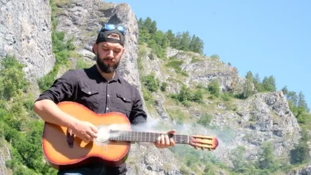Un tipo con barba finge tocar una guitarra acústica de la que sale humo. Extraño video sobre un músico de rock sobre la naturaleza en las montañas
. - Imágenes, Vídeo
