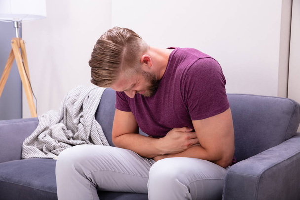 Hombre joven que sufre de dolor de estómago sentado en el sofá en casa
 - Foto, imagen