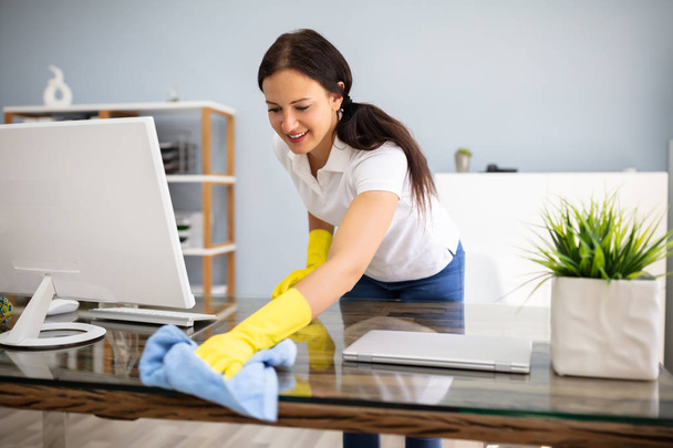 Side View Of Female Janitor Cleaning Desk With Blue Napkin In Office - Foto, Imagen